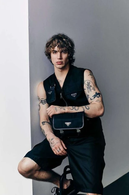 A young adult man with tattoos poses indoors wearing a Prada vest and shorts, holding a Prada bag, under studio lighting against a grey wall.