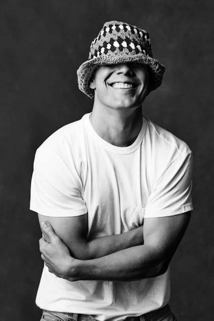 Portrait of a smiling man in a white T-shirt and patterned hat, standing with crossed arms against a grey backdrop in a studio.