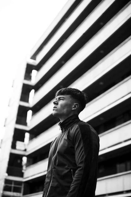 Low-angle portrait in black and white of a young man looking up, wearing a jacket, with a tall building facade in the background.