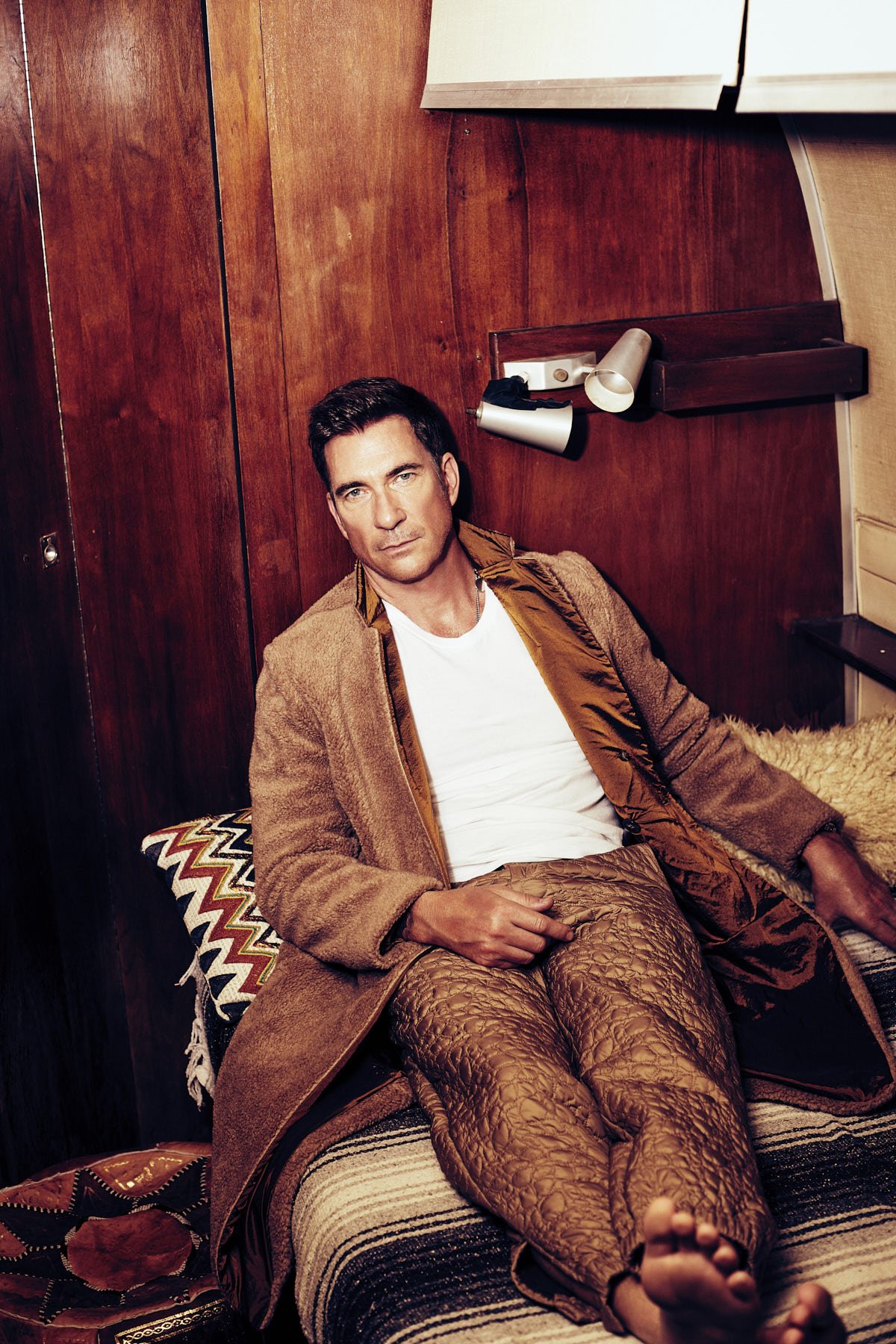 Adult male lounging on a patterned bed in a brown robe and white t-shirt in an indoor setting with wood paneling background.