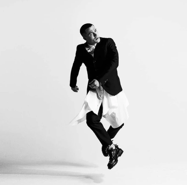 Black-and-white portrait of a man in a suit jacket, dress shirt, and dress pants leaping mid-air with an upward gaze in studio lighting.