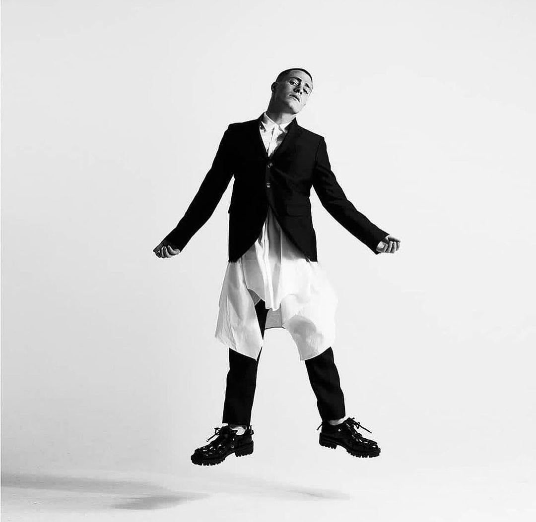 Monochrome full shot of a man in mid-air, wearing a dark blazer, white tunic, black trousers, and shoes against a white backdrop.