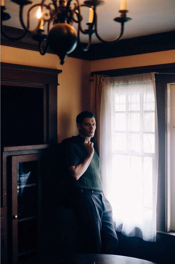 Man in green sweater stands near window with chandelier in background, looking toward the viewer in an indoor setting.