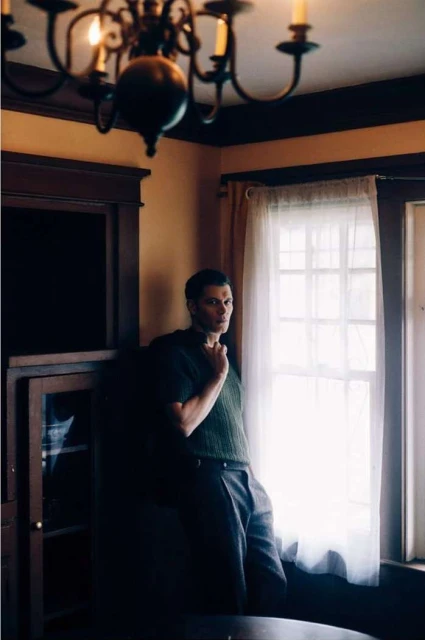 Man in green sweater stands near window with chandelier in background, looking toward the viewer in an indoor setting.
