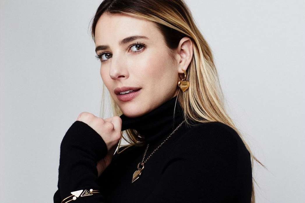 Close-up portrait of a fair-skinned blonde woman wearing a black turtleneck, gold heart earrings, and a gold bracelet, against a white backdrop.
