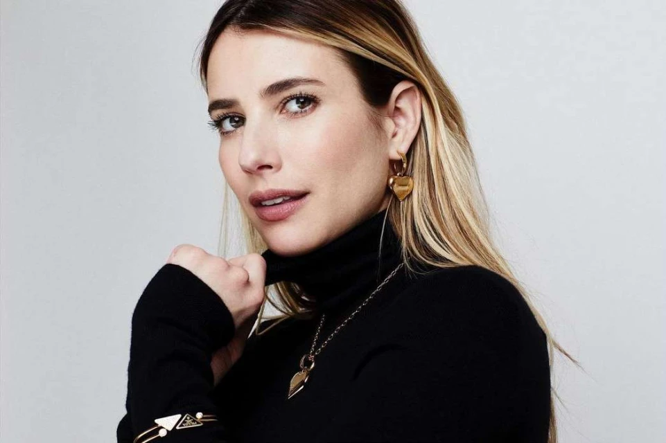 Close-up portrait of a fair-skinned blonde woman wearing a black turtleneck, gold heart earrings, and a gold bracelet, against a white backdrop.