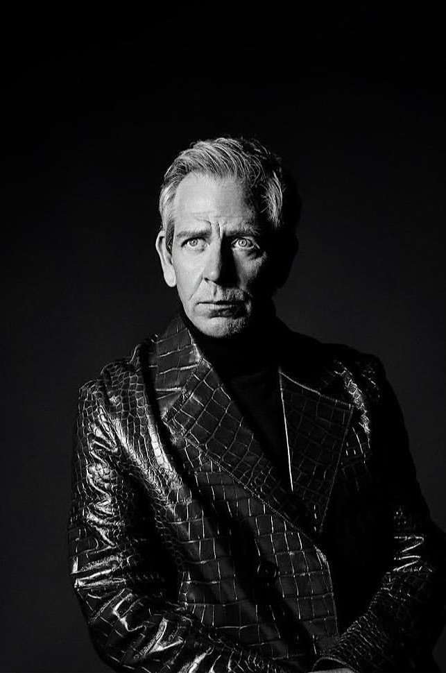Monochrome portrait of a middle-aged man with graying hair, wearing a textured black coat and turtleneck against a dark background.