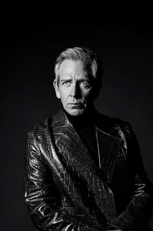 Monochrome portrait of a middle-aged man with graying hair, wearing a textured black coat and turtleneck against a dark background.