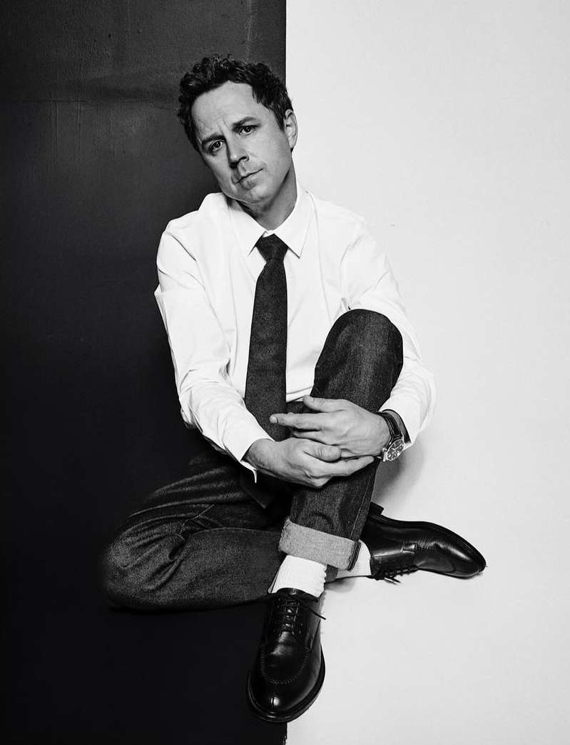 Black and white studio portrait of a man in a white shirt, tie, jeans, and dress shoes, sitting cross-legged on a two-tone background.