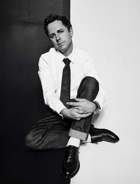 Black and white studio portrait of a man in a white shirt, tie, jeans, and dress shoes, sitting cross-legged on a two-tone background.
