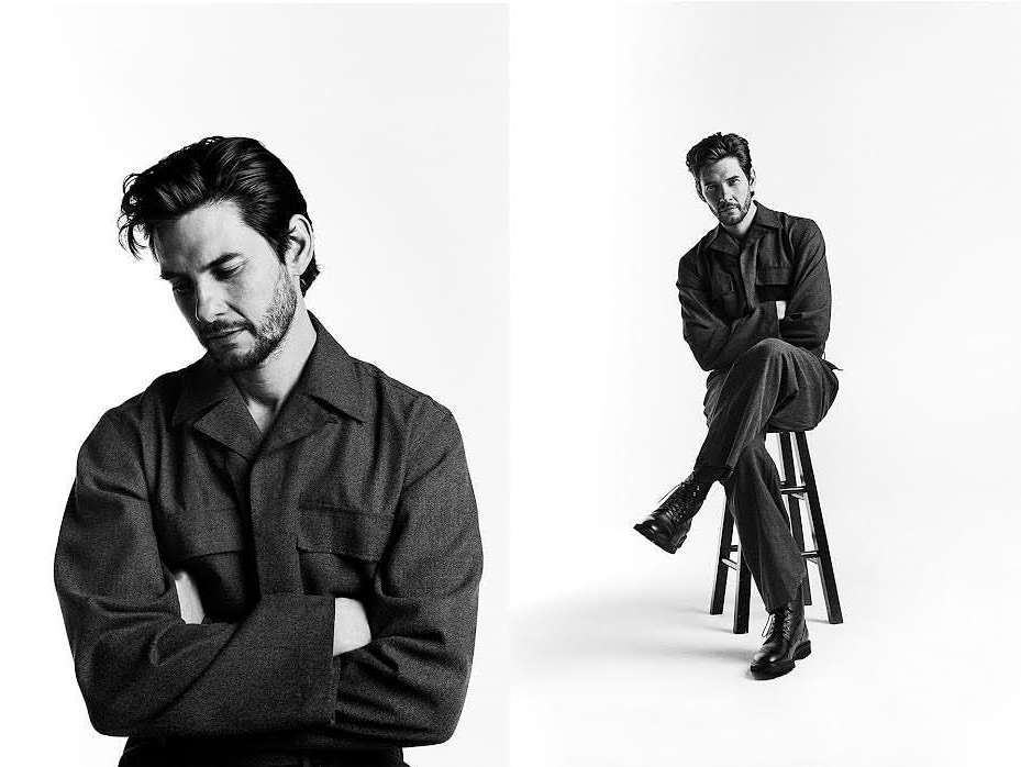 Black-and-white portrait of a man wearing a collared shirt sitting on a stool against a white backdrop in a photo studio setting.