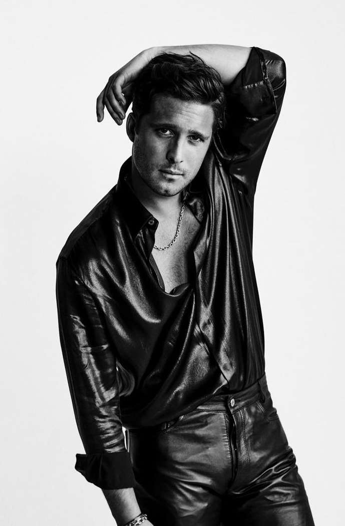 Monochrome portrait of a male model in a black shiny shirt and leather pants, posing with his hand behind his head against a white background.
