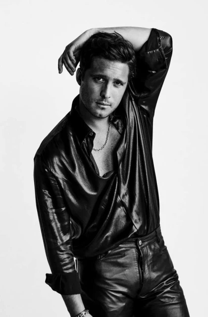 Monochrome portrait of a male model in a black shiny shirt and leather pants, posing with his hand behind his head against a white background.