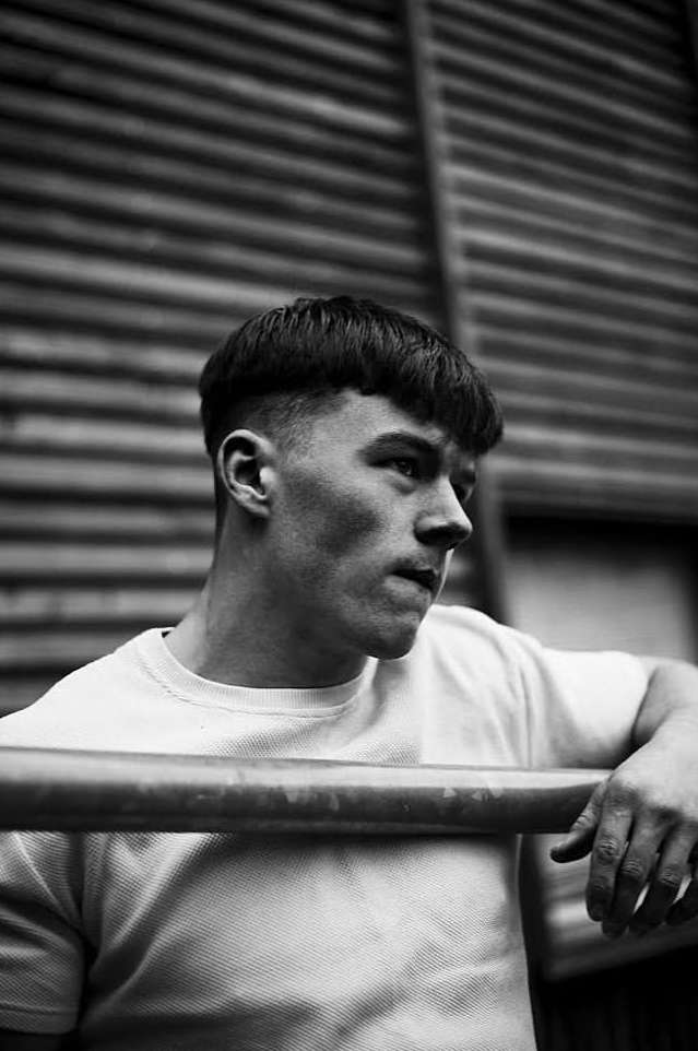 Monochrome portrait of a young adult man with short hair, wearing a light-colored t-shirt, leaning on a metal bar outdoors.