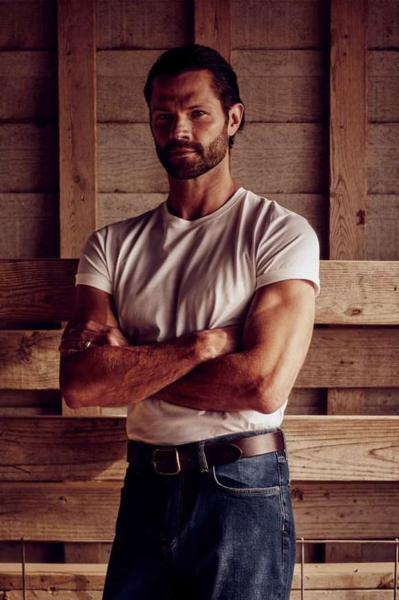 Man with beard and dark hair wearing a white t-shirt and blue jeans standing against a wooden wall