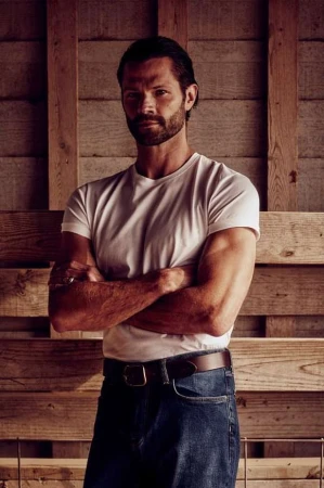 Man with beard and dark hair wearing a white t-shirt and blue jeans standing against a wooden wall