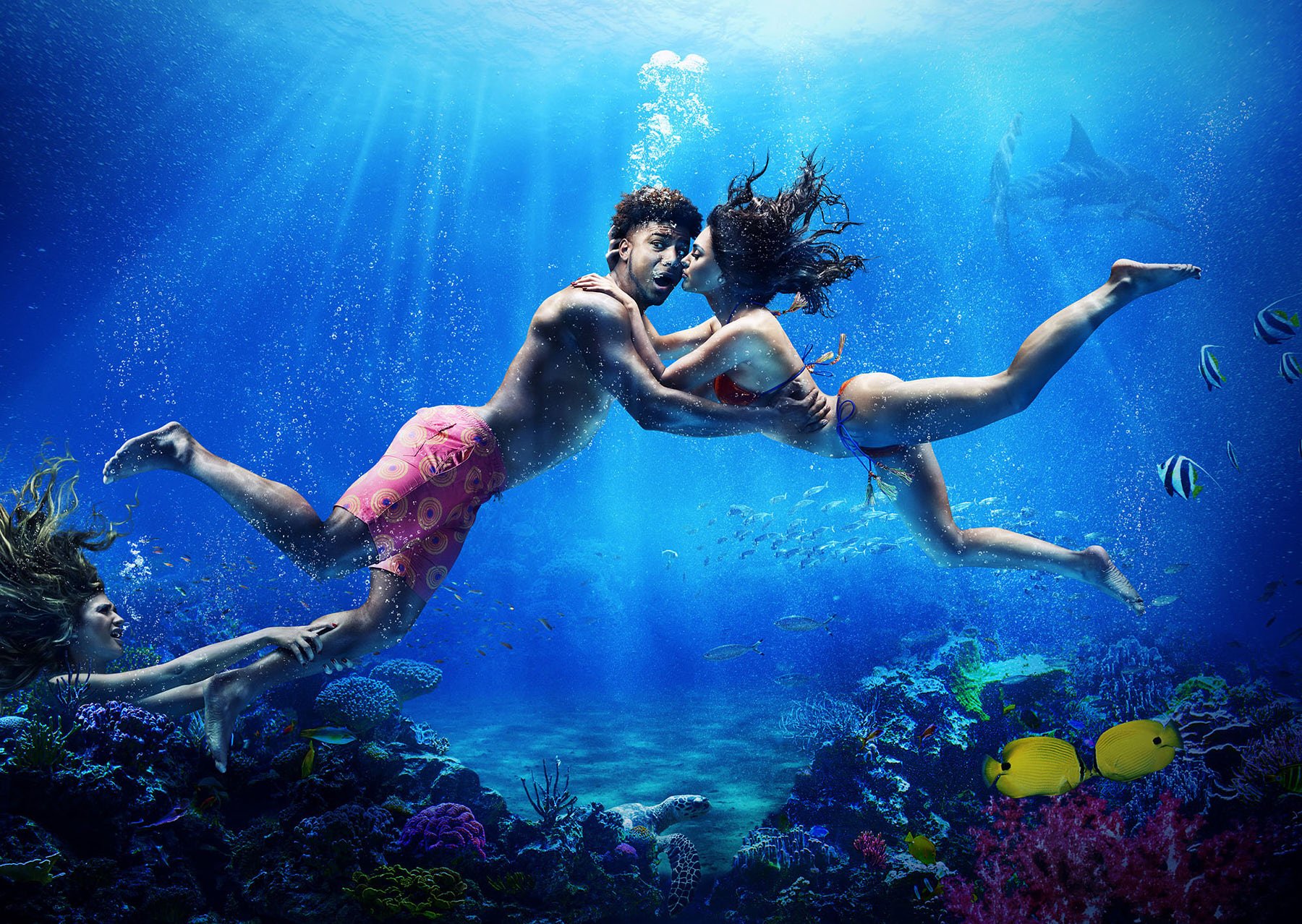 Underwater shot of a couple swimming and embracing among coral reefs and tropical fish, with sunlight streaming from above.