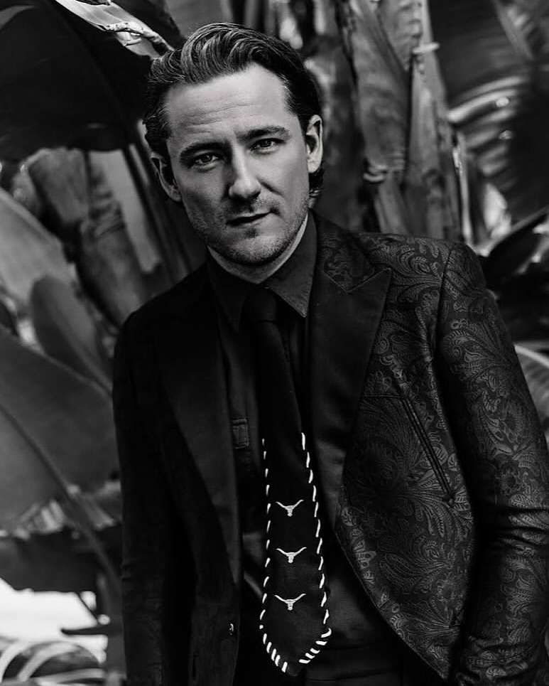 Monochrome portrait of a man in a patterned suit jacket, dark shirt, and tie photographed by Leigh Keily