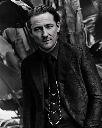 Monochrome portrait of a man in a patterned suit jacket, dark shirt, and tie photographed by Leigh Keily