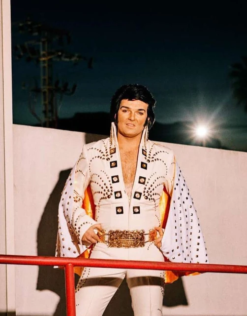 A man in an Elvis Presley jumpsuit stands smiling in front of a white wall and utility pole, illuminated by a nearby light.