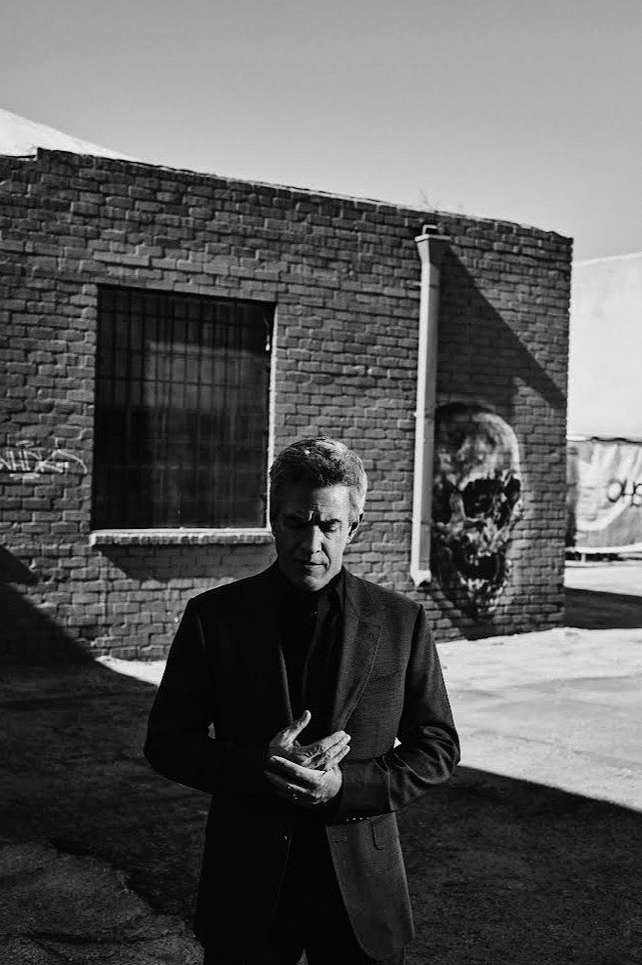 Monochrome portrait of a mature man in a suit standing outdoors against a brick building with a skull mural in soft, natural light.
