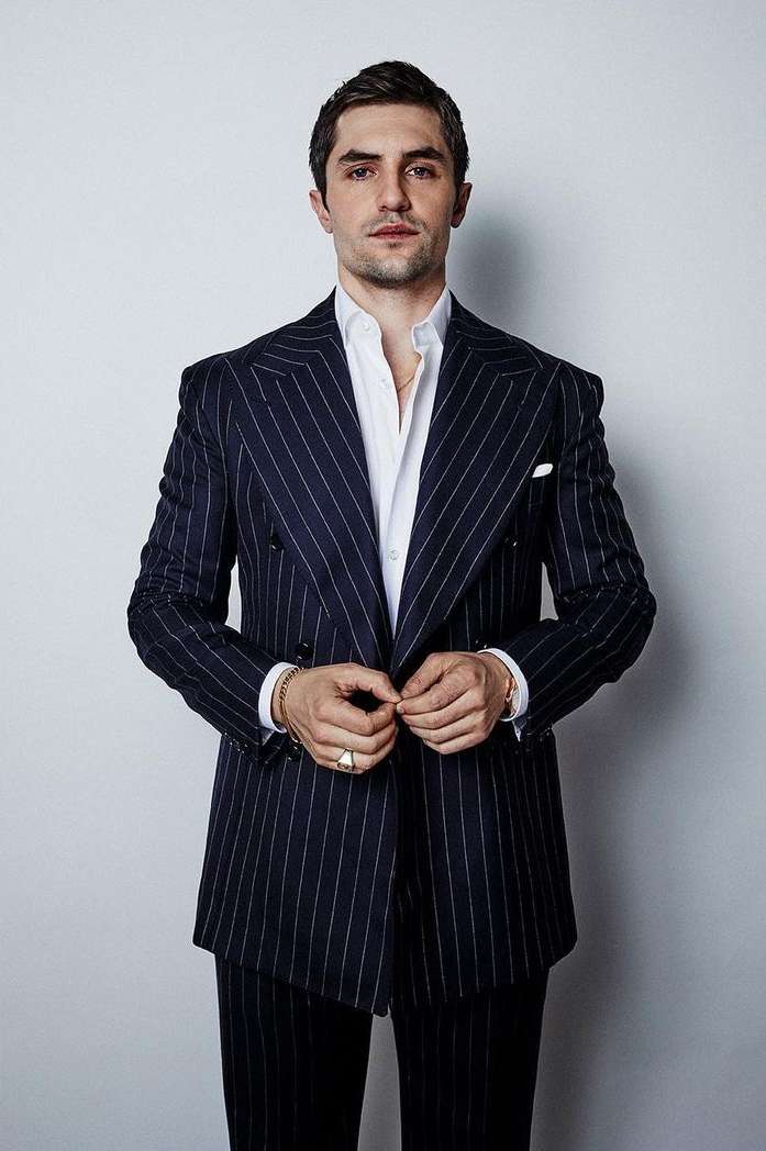 Portrait of adult male in dark pinstripe suit and white shirt, buttoning his jacket, against a plain light background in studio light.