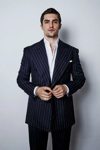 Portrait of adult male in dark pinstripe suit and white shirt, buttoning his jacket, against a plain light background in studio light.