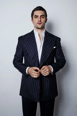 Portrait of adult male in dark pinstripe suit and white shirt, buttoning his jacket, against a plain light background in studio light.