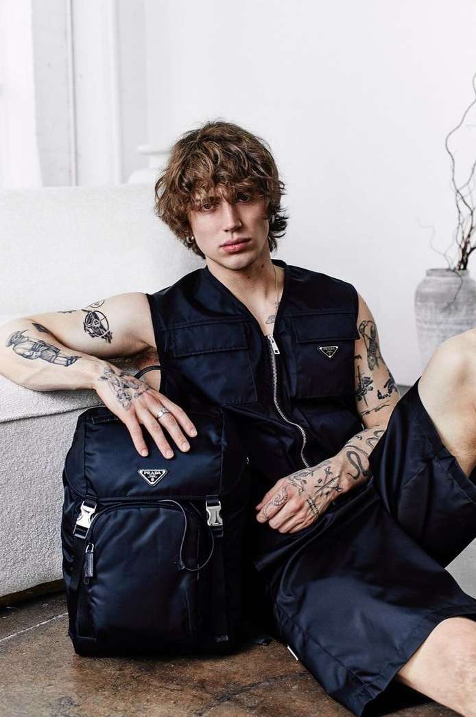 Male model poses with a Prada backpack and vest in a relaxed indoor setting, showcasing fashion and style.