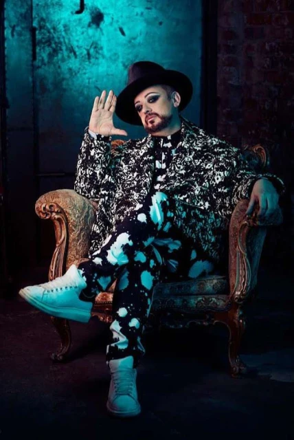 Full-length shot of a man in a black hat and patterned suit sitting in an ornate chair, with teal walls in the background.