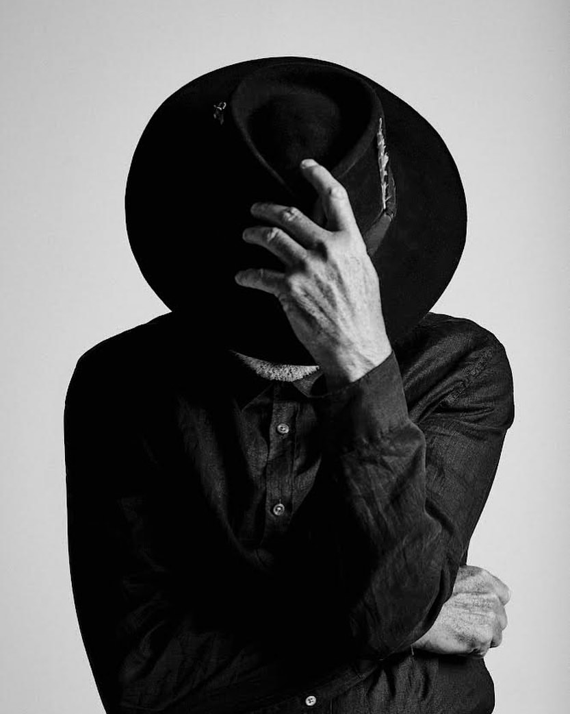 Monochrome portrait of a person wearing a black hat tipped down and a dark buttoned shirt against a plain light background.