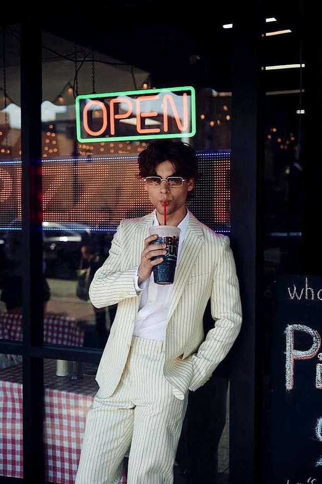 Young adult in a striped suit stands by a pizza restaurant window with an OPEN neon sign, drinking from a large cup through a straw.