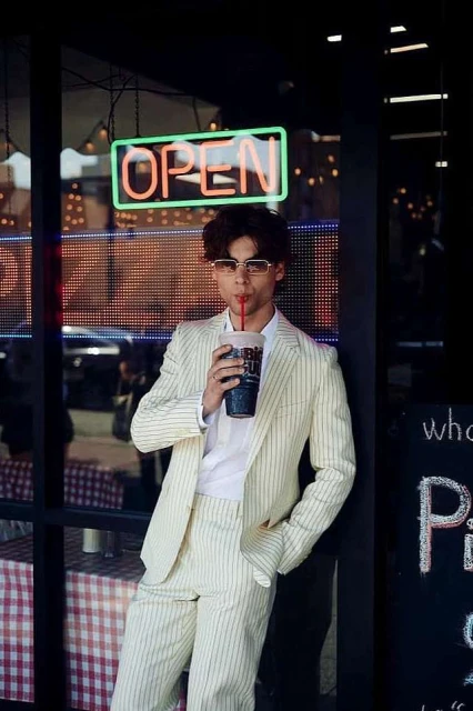 Young adult in a striped suit stands by a pizza restaurant window with an OPEN neon sign, drinking from a large cup through a straw.