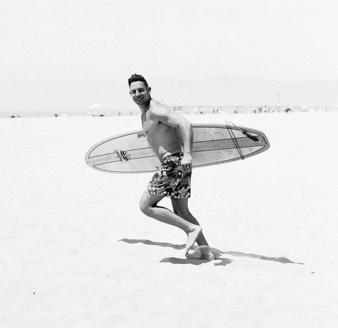 Black and white photograph of a man running across a sandy beach carrying a surfboard under his arm.