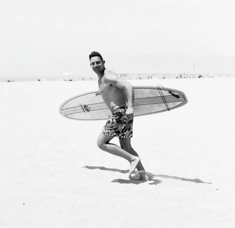 Black and white photograph of a man running across a sandy beach carrying a surfboard under his arm.
