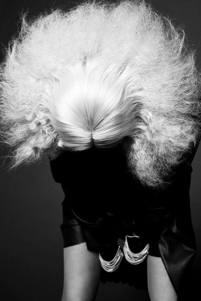 Black and white fashion portrait of a person with a large blonde afro hairstyle, wearing a black top and statement silver earrings.
