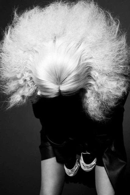 Black and white fashion portrait of a person with a large blonde afro hairstyle, wearing a black top and statement silver earrings.