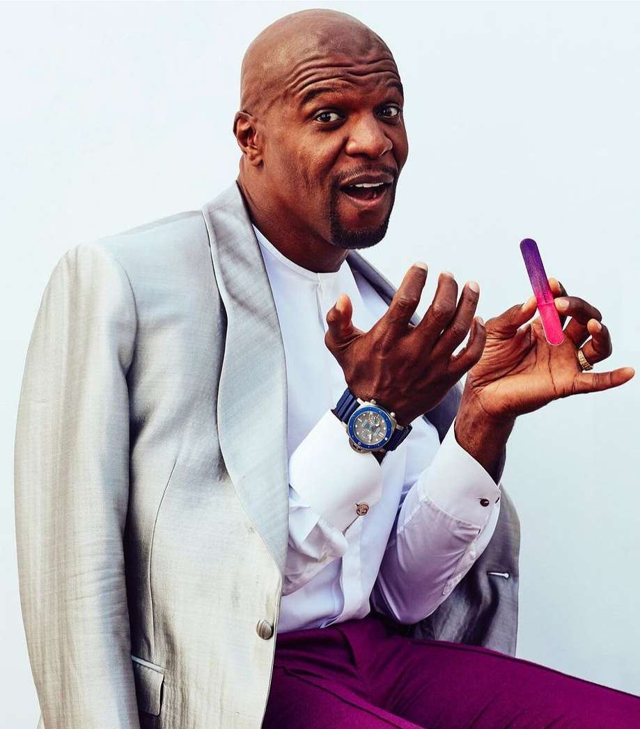 Bald adult male in grey jacket holds pink and purple nail file indoors, smiling with his mouth open in a brightly lit setting.