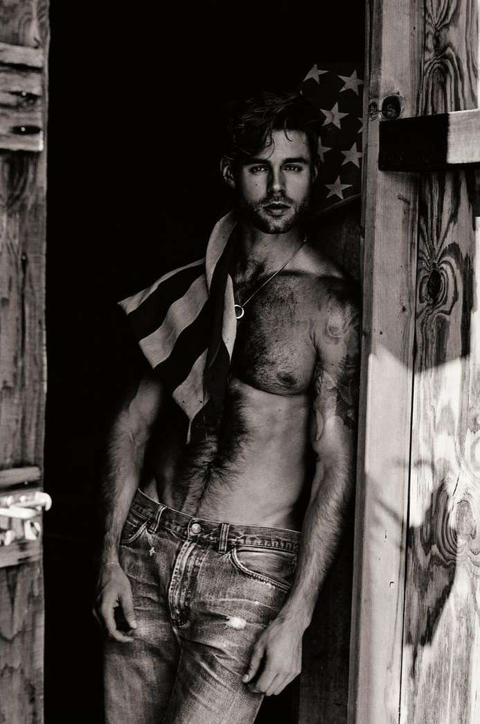 Black and white portrait of a shirtless man wearing jeans, leaning against a wooden door with an American flag draped over his shoulder.