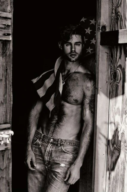 Black and white portrait of a shirtless man wearing jeans, leaning against a wooden door with an American flag draped over his shoulder.