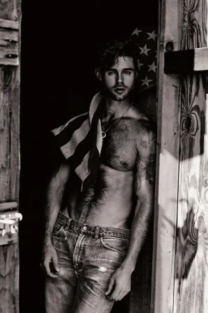 Black and white portrait of a shirtless man wearing jeans, leaning against a wooden door with an American flag draped over his shoulder.