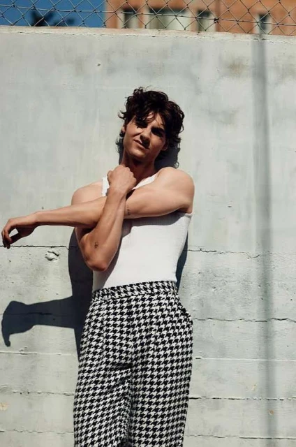 A casually dressed adult male leans against a textured concrete wall outdoors, wearing a white tank top and black and white houndstooth pants.