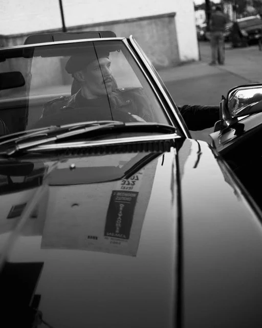 Black and white shot of an adult male in a convertible with a building and another person in the blurred background.