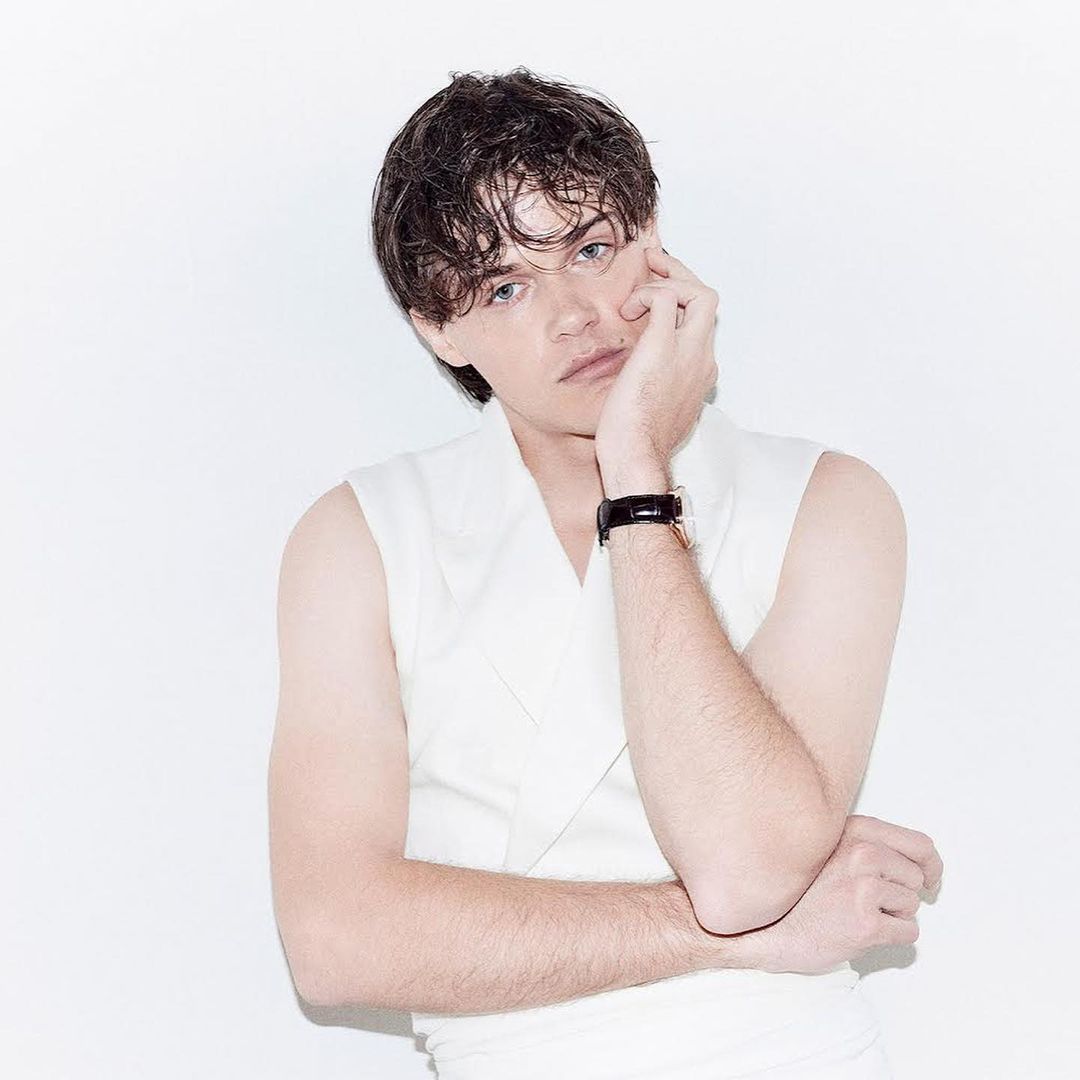 Portrait of a young adult male with dark hair wearing a sleeveless white top, posing with his hand near his face against a white backdrop.