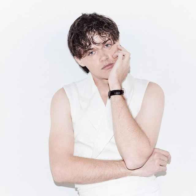 Portrait of a young adult male with dark hair wearing a sleeveless white top, posing with his hand near his face against a white backdrop.