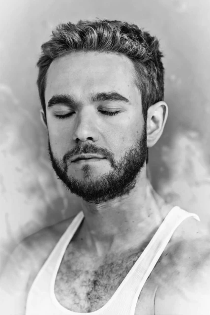 Black and white portrait of a man with a beard, closed eyes, and a white tank top against a textured gray and white backdrop.