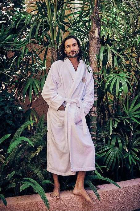 Adult man in a white robe standing outdoors against a backdrop of tropical greenery and palm trees, with natural light.