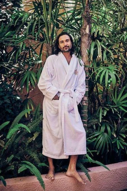 Adult man in a white robe standing outdoors against a backdrop of tropical greenery and palm trees, with natural light.