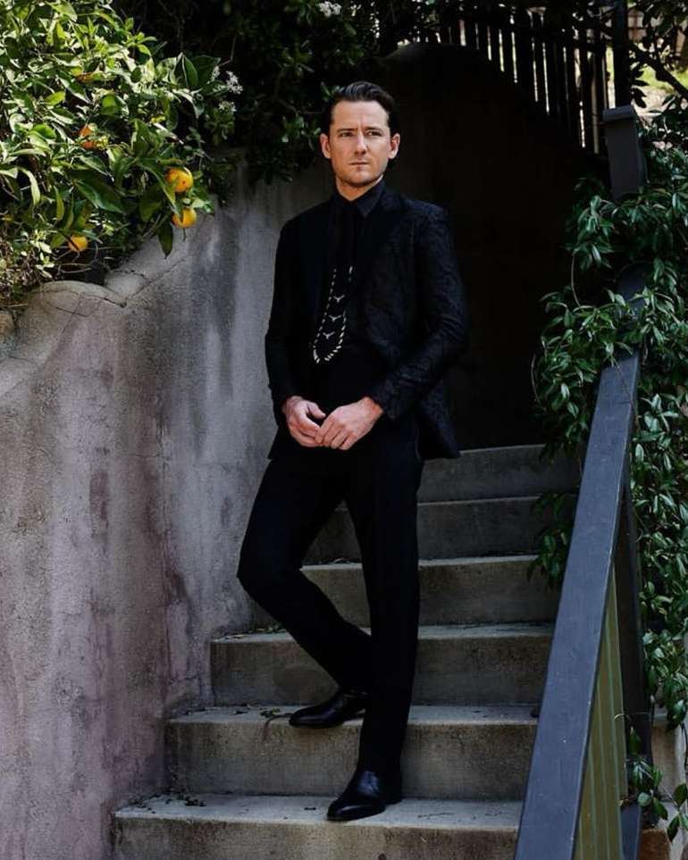 A man in a black suit stands on stone steps surrounded by greenery. He is looking off camera in a portrait view with outdoor setting.