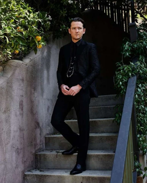 A man in a black suit stands on stone steps surrounded by greenery. He is looking off camera in a portrait view with outdoor setting.